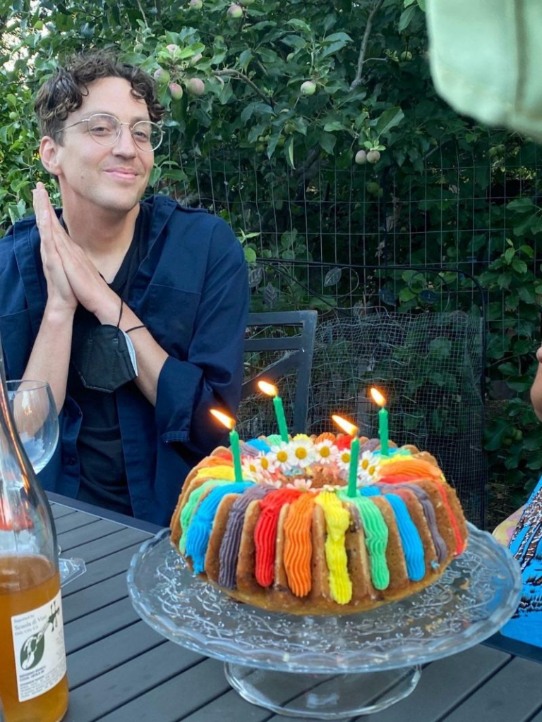 David Imhoff smiles into the camera. Dark, curly, medium length hair. Glasses. Hands pressed together. Navy blue shirt. Trees in background. Rainbow poundcake in foreground with 4 green lit candles.